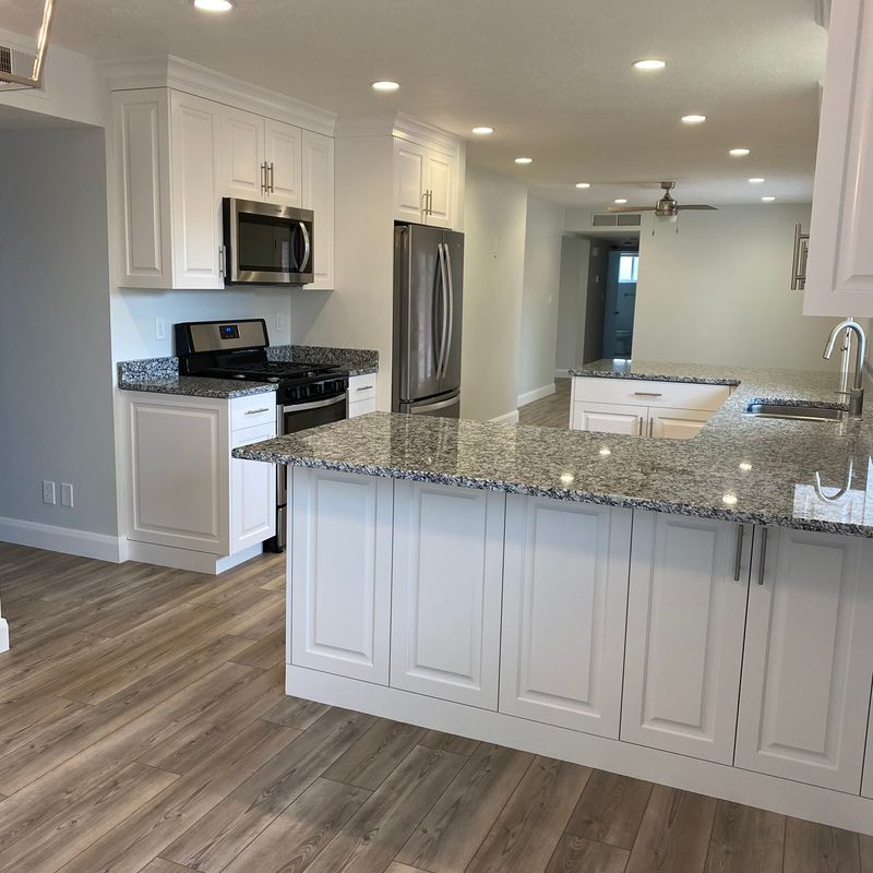 Modern white kitchen cabinets with a gray granite countertop extending into an island, stainless steel appliances, and wood-look flooring.