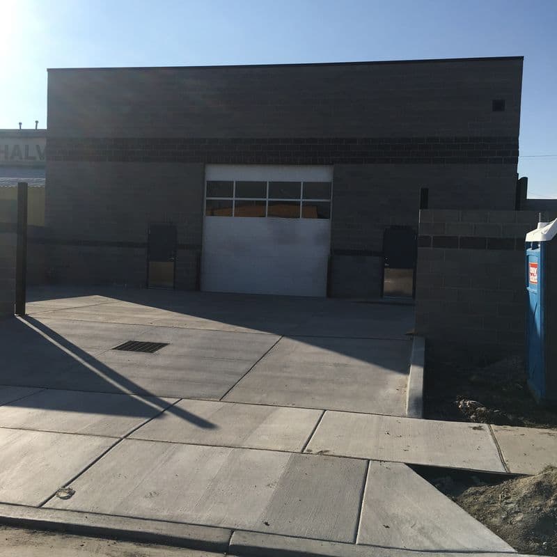 Exterior of a gray concrete auto repair garage with a white lifting door, on a sidewalk with concrete squares.