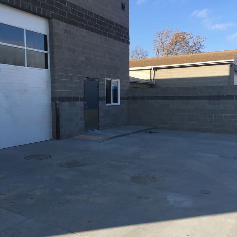 Exterior of a modern building with concrete brick walls and a white lifting door, under a blue sky with a leafless tree in the background.