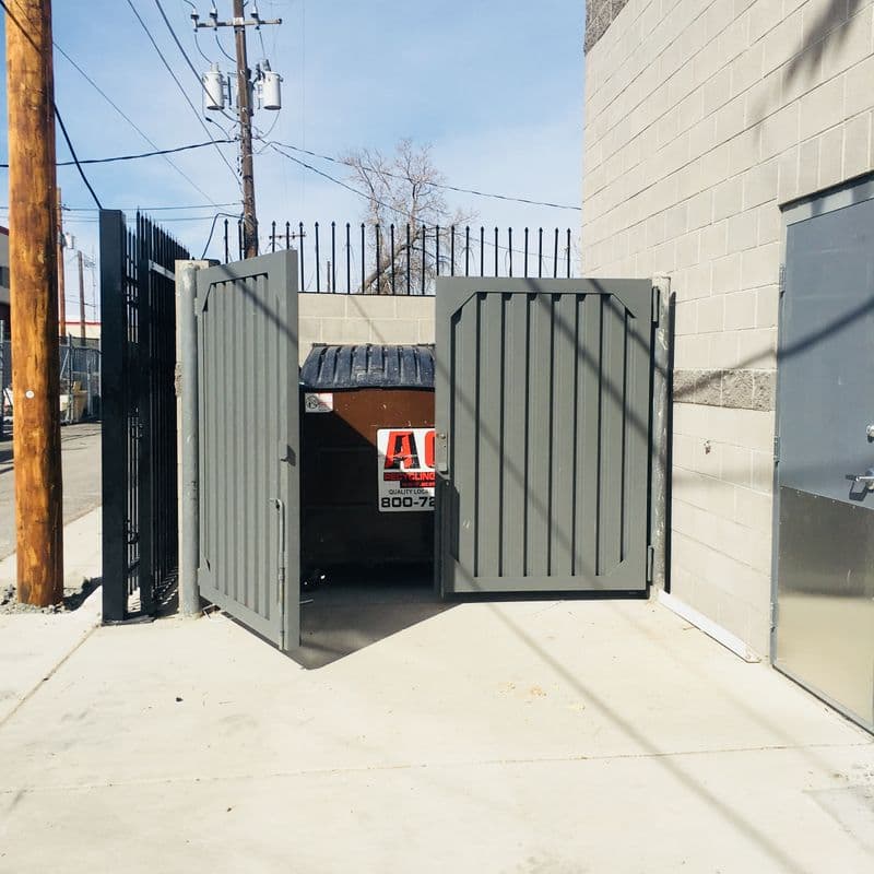 An olive-green enclosure partially covering a large commercial dumpster.